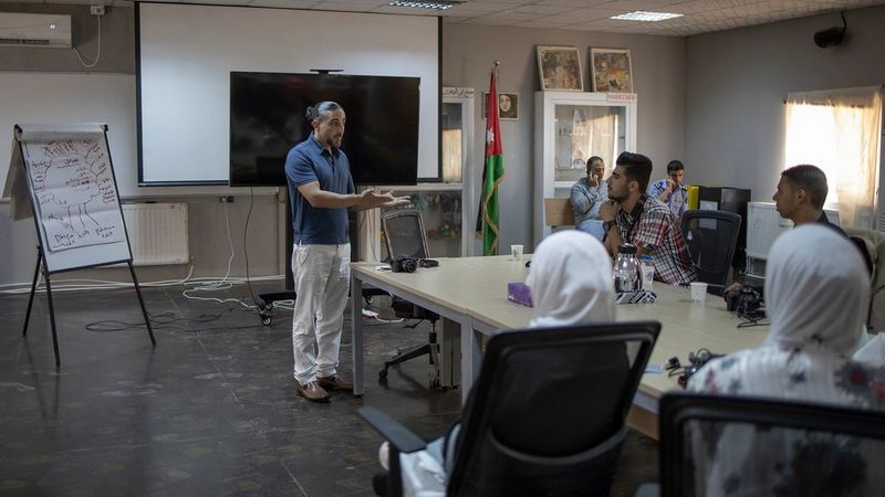 Muhammed Muheisen, wearing white trousers and a blue polo shirt, stands in a classroom, in front of a whiteboard and beside a flip chart. He gestures with both hands outstretched in front of him to a table of students.