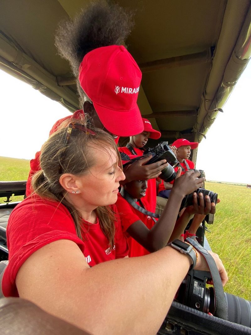 A woman and a man, both dressed in red t-shirts, are sat in the back of a truck on a game drive. They look into the viewfinder of a camera.