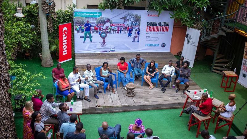 A group of people sitting on a wooden platform on blue chairs in front of an audience. Behind them is a Canon banner and a huge banner saying ‘Canon Alumni Exhibition’ in front of it. They are all surrounded by grass, trees and foliage.