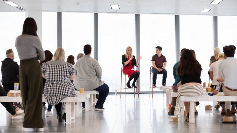 A woman in a red dress sits on a high stool and speaks to a group of people in a bright white room.