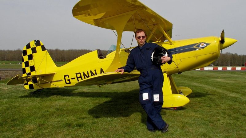 A man in a flight suit and sunglasses stands in front of a small yellow light aircraft.