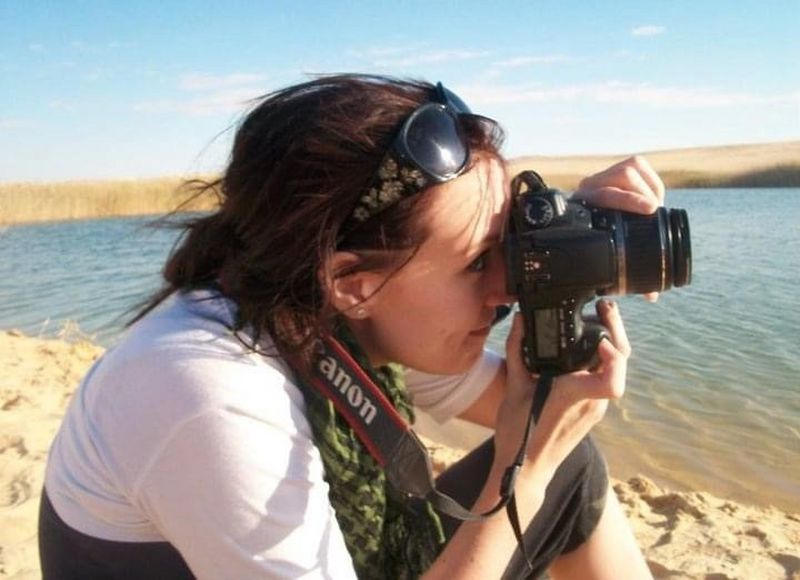A woman crouching on a beach holds a Canon camera to her face.