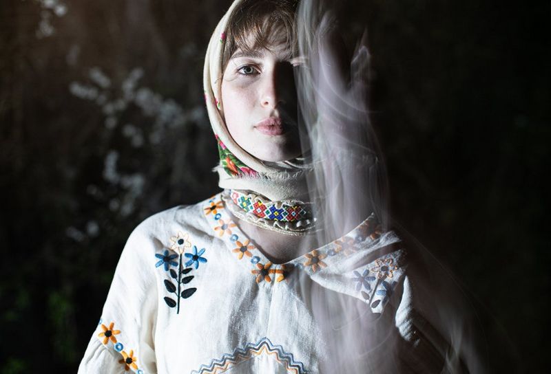 A young woman stands, wearing traditional Ukrainian dress. In front of her to the right is a ghostly figure of herself, representing her spirit.