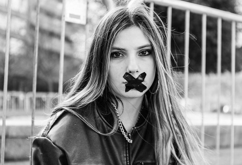 A black and white portrait of a young woman with long blonde hair. She has black tape across her mouth in an X.