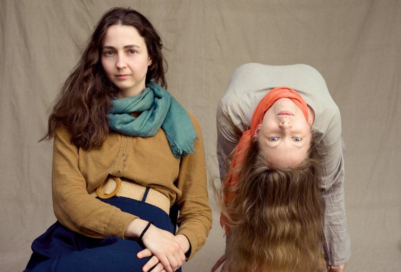 On the left, a woman sits facing the camera. On the right, another woman’s body faces the wall behind, but she bends backwards so that her upside-down face can be seen and hair falls to the ground.