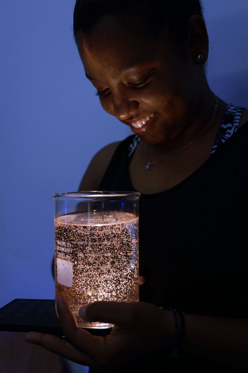 A person smiles in the dark while holding a large glass beaker filled with glowing, suspended gametes.