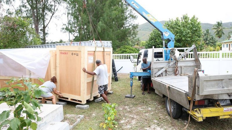 Workers use a crane truck to move large wooden crates marked ‘Fragile’ in an outdoor tropical
