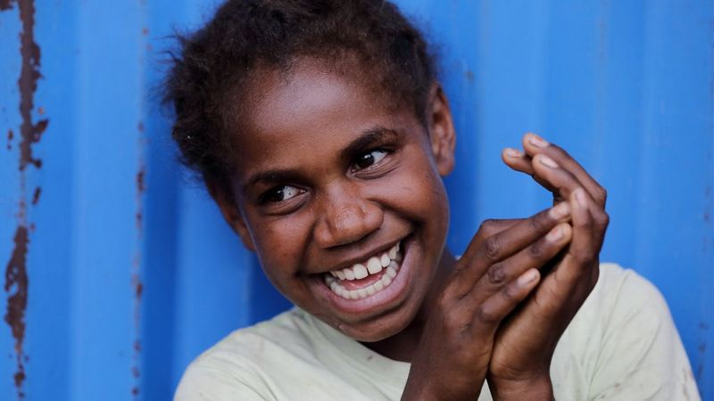 A young girl of colour stands in front of a wall of blue corrugated metal. She has a big smile and is clasping her hands together in delight