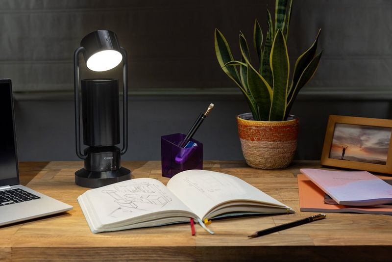 Een Canon Speakerlamp ML-A staat op een houten bureau naast een boek, een plant en wat kantoorartikelen.