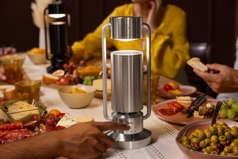 Twee Canon Speakerlampen ML-A staan op een volle eettafel, omgeven door borden met eten en mensen die op stoelen zitten. Een hand reikt uit en past de bedieningsknoppen aan van de dichtstbijzijnde Speakerlamp ML-A.