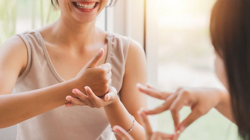 A woman from the chin down, the camera focuses on her hands, which are engaged in sign language. The hands and hair of the person she is communicating with are blurred and slightly out of the image, but their hands are also signing.  