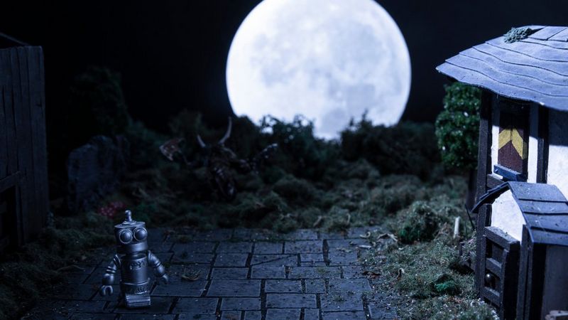 A nightime scene with a huge silver moon as a backdrop. To the right is a small shack-type house with a sloped roof and a single window. To the right and centre, is a tree trunk and there is foliage in front of the moon. In the front left shadows stands a small silver robot.
