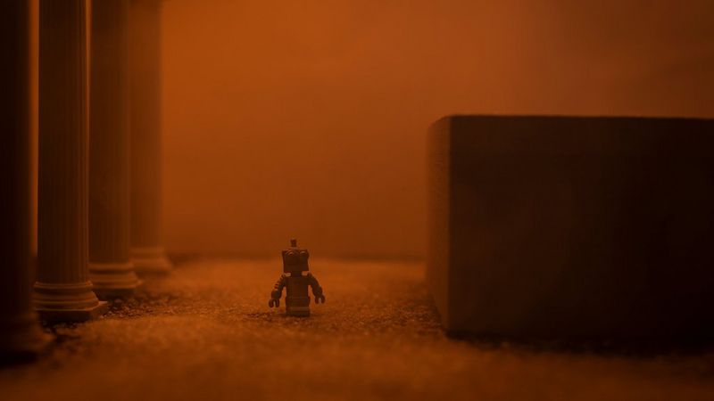 A tiny robot stands between a large granite-looking block on the right and four decorative columns on the left. The scene has a dusty, red orange glow.