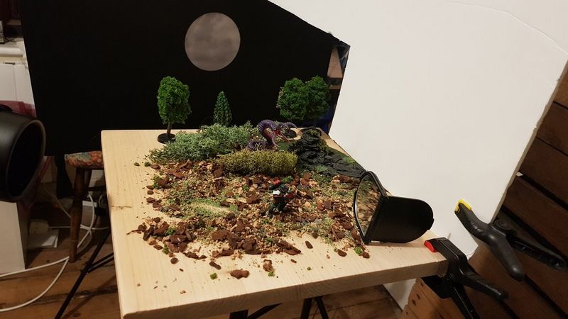 Another behind the scenes shot. This time the table is viewed from the right-hand side and has a white backdrop clipped to it. Again, there is soil and moss, but also miniature trees.