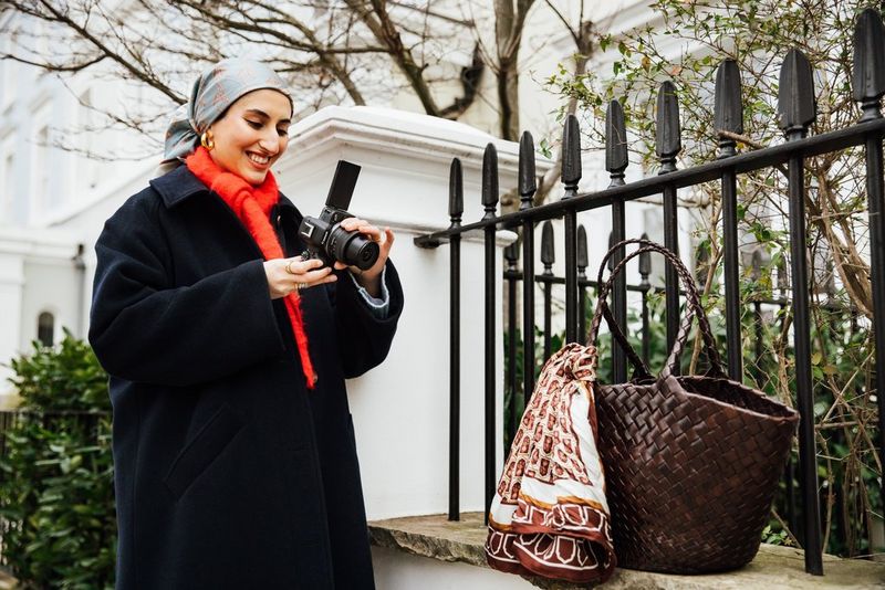 A person points a Canon PowerShot V1 at a large woven basket with a scarf around one handle resting against iron railings, with trees and buildings in the background.