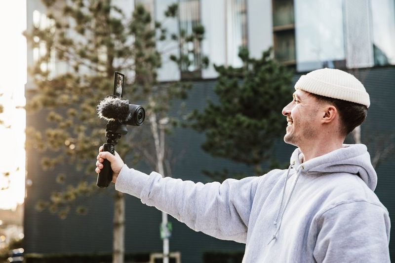 A smiling man on a city street holds a Canon EOS R50 V on a grip at arm's length.