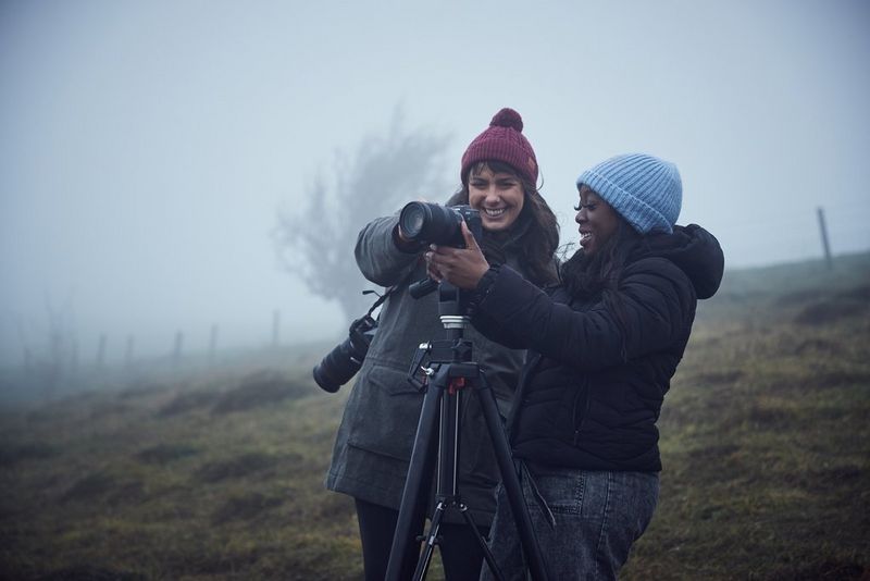 Two women, both wearing winter clothing, on a foggy hillside, adjusting a Canon camera on a tripod.