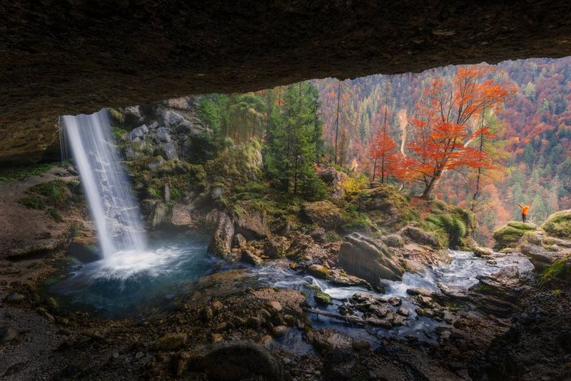 Iemand staat aan de rand van een klif en kijkt uit over een vallei met bomen, met links op de voorgrond een waterval. Foto gemaakt door Luka Vunduk met de Canon EOS R5 Mark II.