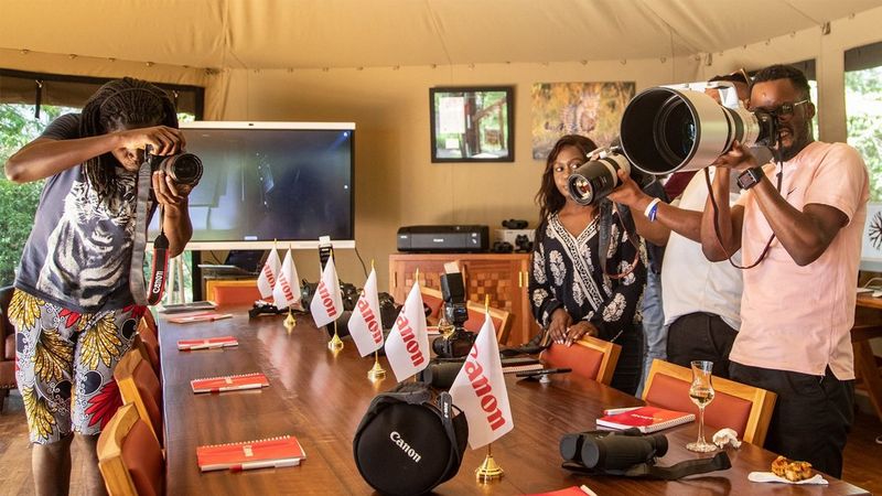A brown boardroom table has six Canon flags spaced across its length. It is in what looks to be a large white marquee-like construction. Around the table, three people test out Canon cameras, while a further two look on. © Usha Harish