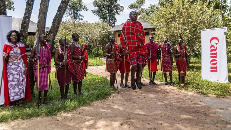 Ten men and women wearing the traditional dress of the Maasai tribe stand on a grassy verge next to a pop-up banner displaying the Canon logo. The man in the centre of the image jumps high into the air. © Peter Ndungu