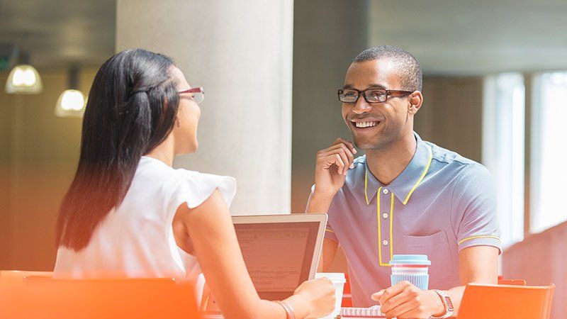 Man and woman sat opposite on work table with laptop open and coffee, in casual meeting