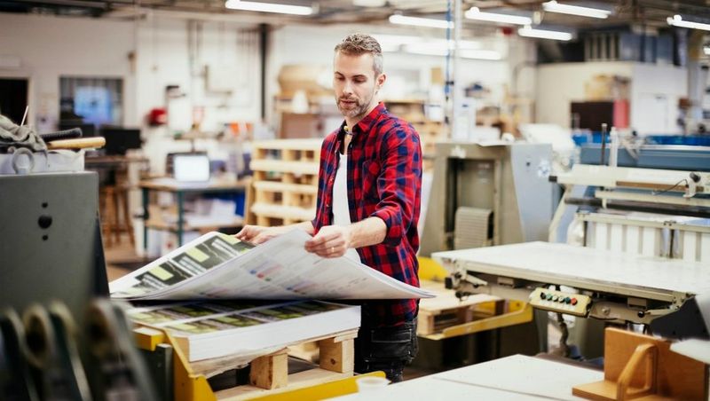 Girl in red check shirt and man in blue t-shirt working at a digital printer with colour charts and ink on the back wall