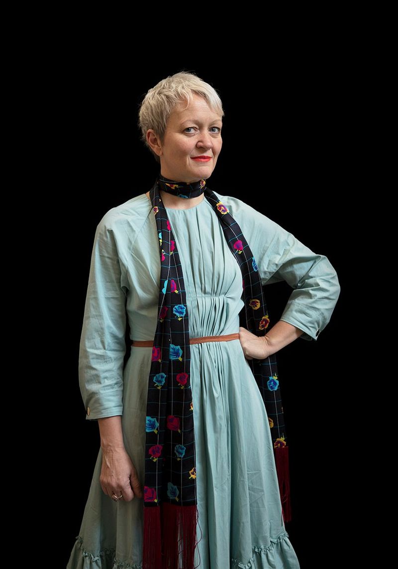 A woman with short blonde hair wears a green dress and scarf and stands against a black background.