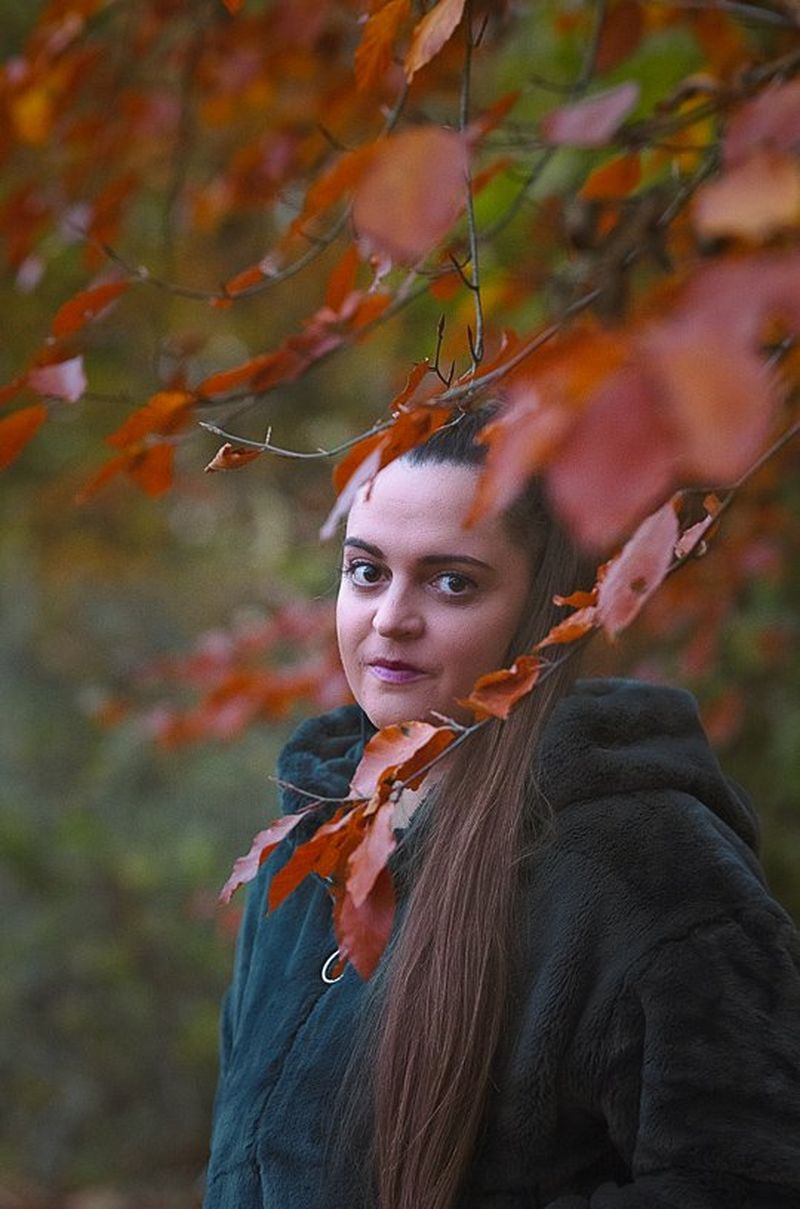 A woman in a black halter-neck dress stands by foliage. She has long dark curly hair.