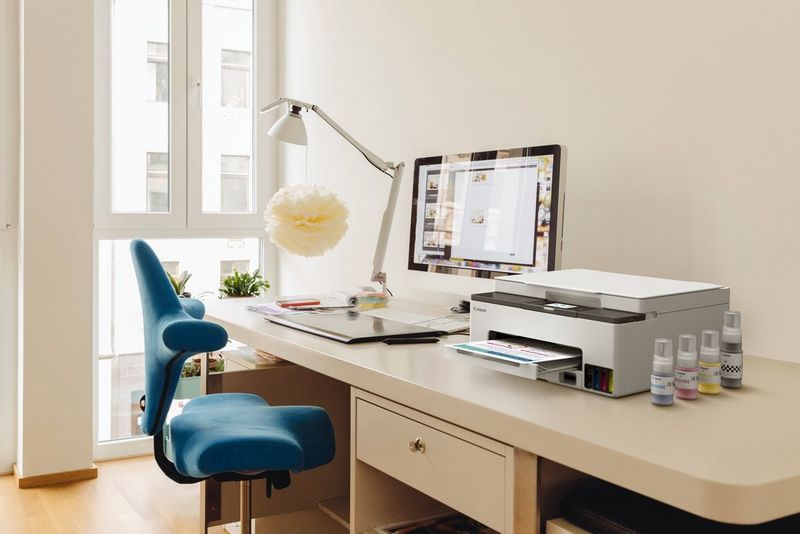 Принтер Canon «Формула выгодной печати» и несколько контейнеров с чернилами лежат на столе перед монитором и настольной лампой.