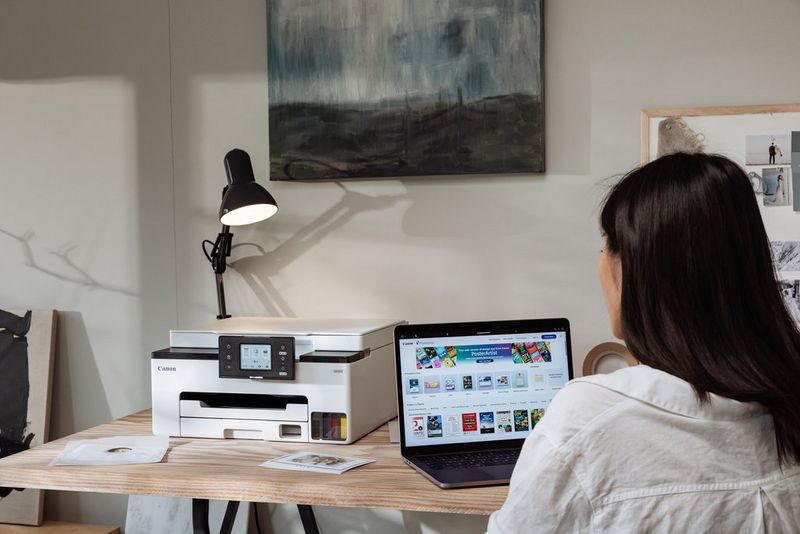 A woman sits at a laptop at a wooden desk. Next to her on the worktop is a Canon MAXIFY printer.