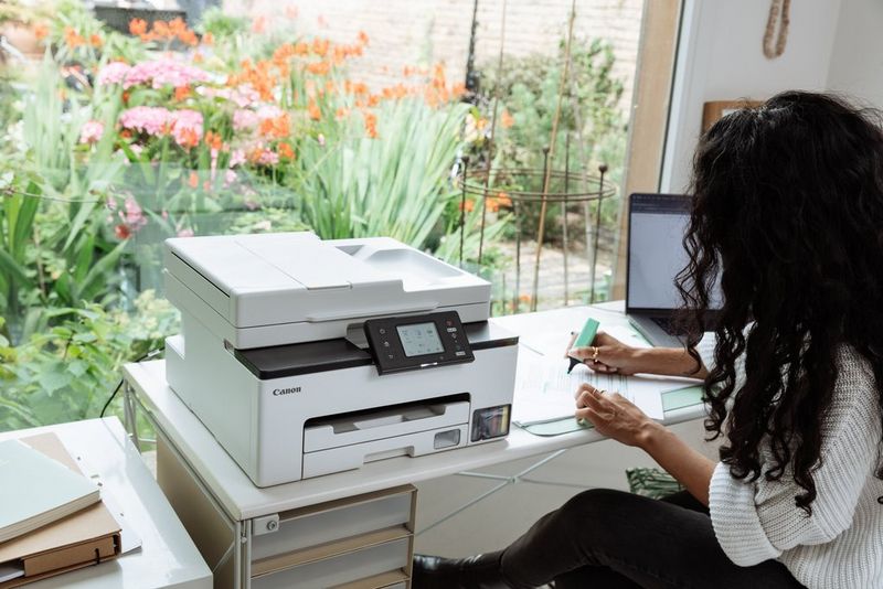 אישה מדגישה טקסט על פיסת נייר המונח על שולחן, לידה מדפסת Canon MAXIFY GX וגן שופע שנשקף מבעד לחלון גדול מאחורי השולחן.