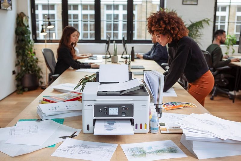 מדפסת הזרקת דיו Canon MAXIFY GX למילוי חוזר מונחת על שולחן גדול במשרד, כשמסביבה ערימות של מסמכים ואנשים עובדים ברקע.