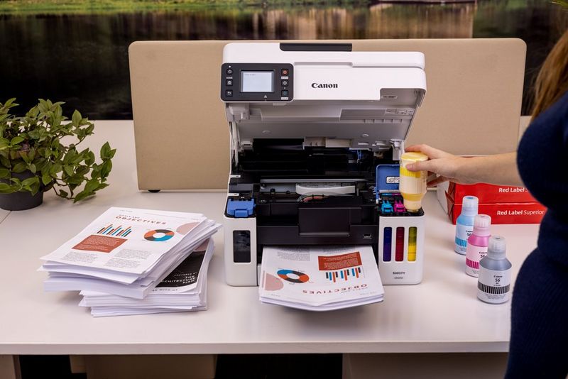 A user refills the yellow ink in a Canon MAXIFY MegaTank printer, the other three CMYK colours on the table beside, along with stacks of printed documents.