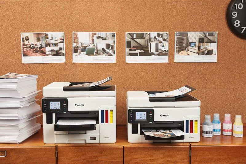 Een MAXIFY GX7050 en MAXIFY GX6050 op een werkblad naast een grote stapel papier en printerinkt.