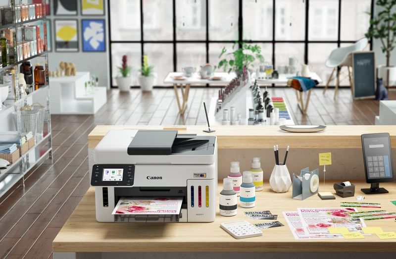 מדפסת Canon MegaTank מונחת על שולחן עץ בסטודיו מודרני ומרווח, כשברקע חלונות בגובה מלא.