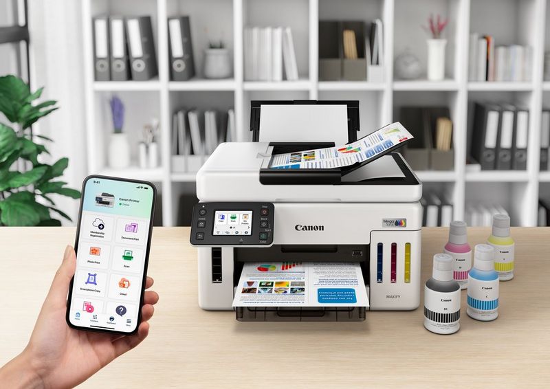 משתמש מחזיק סמארטפון שבו מופיעה אפליקציית Canon PRINT ששולטת במדפסת הזרקת דיו Canon MAXIFY למילוי חוזר. המדפסת מונחת על שולחן ולצידה בקבוקי דיו.