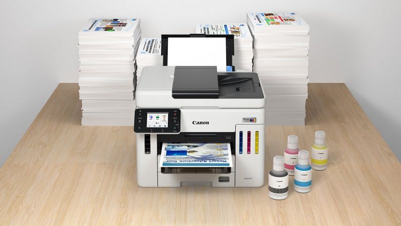מדפסת הזרקת דיו Canon MAXIFY GX למילוי חוזר על שולחן מעץ, עם ערימות גדולות של תדפיסים מאחוריה וארבעה בקבוקי דיו לצידה.