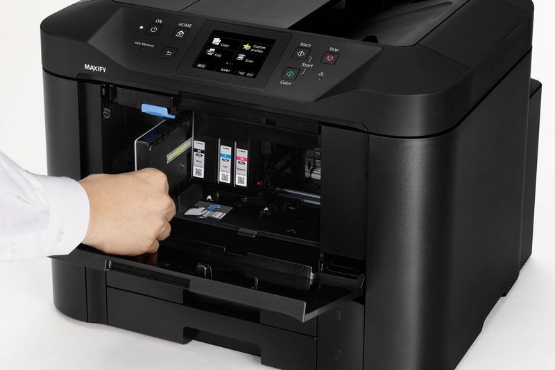 A user's hand removes one ink cartridge from a Canon MAXIFY printer, open at the front.
