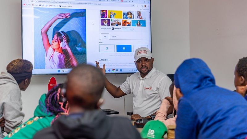 A man in a white Canon-branded shirt and baseball cap gestures towards a large screen showing an image of a young woman in headphones, dancing. Looking at him, with their backs to the camera are several young students.