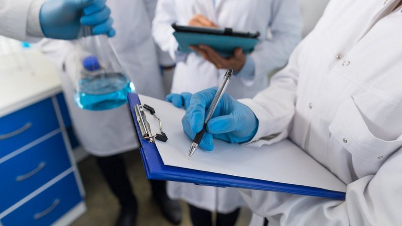 The upper bodies of three clinicians, wearing white lab coats and blue plastic gloves. The one closest to camera holds a clipboard and pen. Another holds a conical flask of blue liquid.