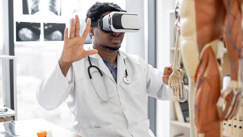 A doctor in a white coat and wearing a VR headset holds his hands up. Beside him is an anatomical skeleton and some scans are stuck to a board behind him.