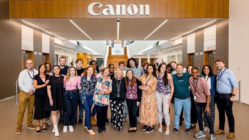 Twenty-five people of all ages, colours, shapes, sizes and clothing, stand in the foyer of Canon’s EMEA and UK HQ. Above them is a huge Canon logo in silver, mounted against a wooden background. Behind them are a set of stairs leading to the first floor of the building. On their left and right are umber coloured walls with doors set into them. The floor is tiles and a light brown in colour. (Image courtesy of CG Weddings)