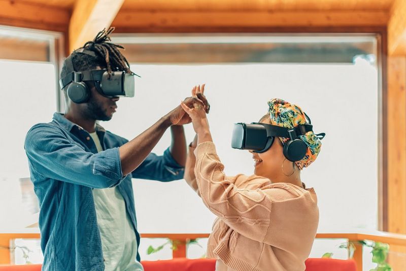 On the left stands a man in a blue shirt over a white t-shirt. He is wearing a black VR headset and his locs are falling over the top of it. His arms are raised in front of him and he hold hands with a woman on the right of the photograph. She is also wearing a VR headset over a colourful headscarf and a peach-coloured chunky knit sweater.