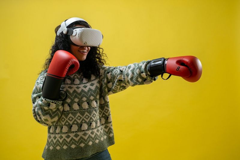 A smiling woman stands, slightly to one side against a yellow background. She has shoulder length dark curly hair. She is wearing a green and white patterned winter jumper and jeans, and a white VR headset with connected red boxing gloves. She has her right glove near her face and left arm outstretched, suggesting that she is boxing in a virtual reality world.