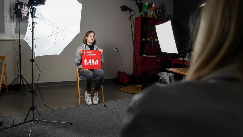 A girl in glasses sits on a wooden chair, holding a red sign showing the 1st United Nations Sustainable Development Goal, written in Polish. She has studio lights shining on her and to the right of the image is the close up of the back of another student, who is presumably taking a photograph of her.