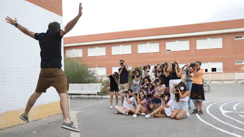 A man in brown shorts and a black t-shirt faces a crowd of young people, who are all holding cameras to their faces to take his photograph. He is leaping in the air, doing a star jump.