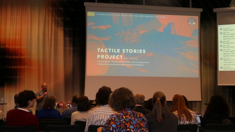 Un uomo parla di fronte a un pubblico e davanti a uno schermo su cui compare la scritta "TACTILE STORIES PROJECT".
