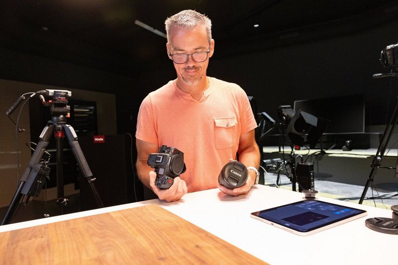 Мужчина в оранжевой футболке держит в руках беззеркальную камеру Canon с объективом RF, а рядом с ним на столе лежит цифровой планшет.