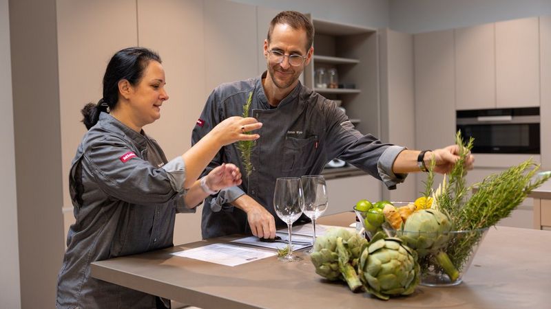 Мужчина и женщина стоят на кухне в серой форме шеф-поваров с логотипом Miele, держа в руках зелень и травы. Перед ними стоят два пустых бокала для вина, а также стеклянная емкость с сырыми овощами, включая артишоки.