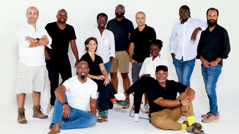 11 people posing for the camera against a white background, some seated on the floor and others are stood.